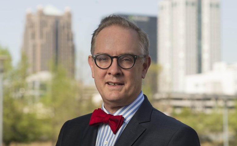 Dr. Barry Sleckman, Cancer Center Director, in a suit and bowtie, with the Birmingham skyline behind him, representing UAB’s leadership in oncologist recruitment.