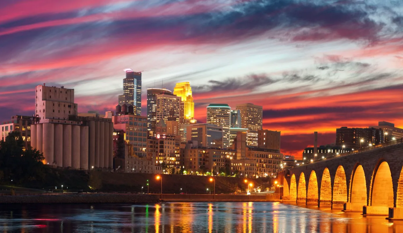 A stunning sunset over downtown Minneapolis, featuring the illuminated skyline and the historic Stone Arch Bridge reflecting on the Mississippi River—home to the University of Minnesota Medical School and its distinguished Chair of Neurosurgery.