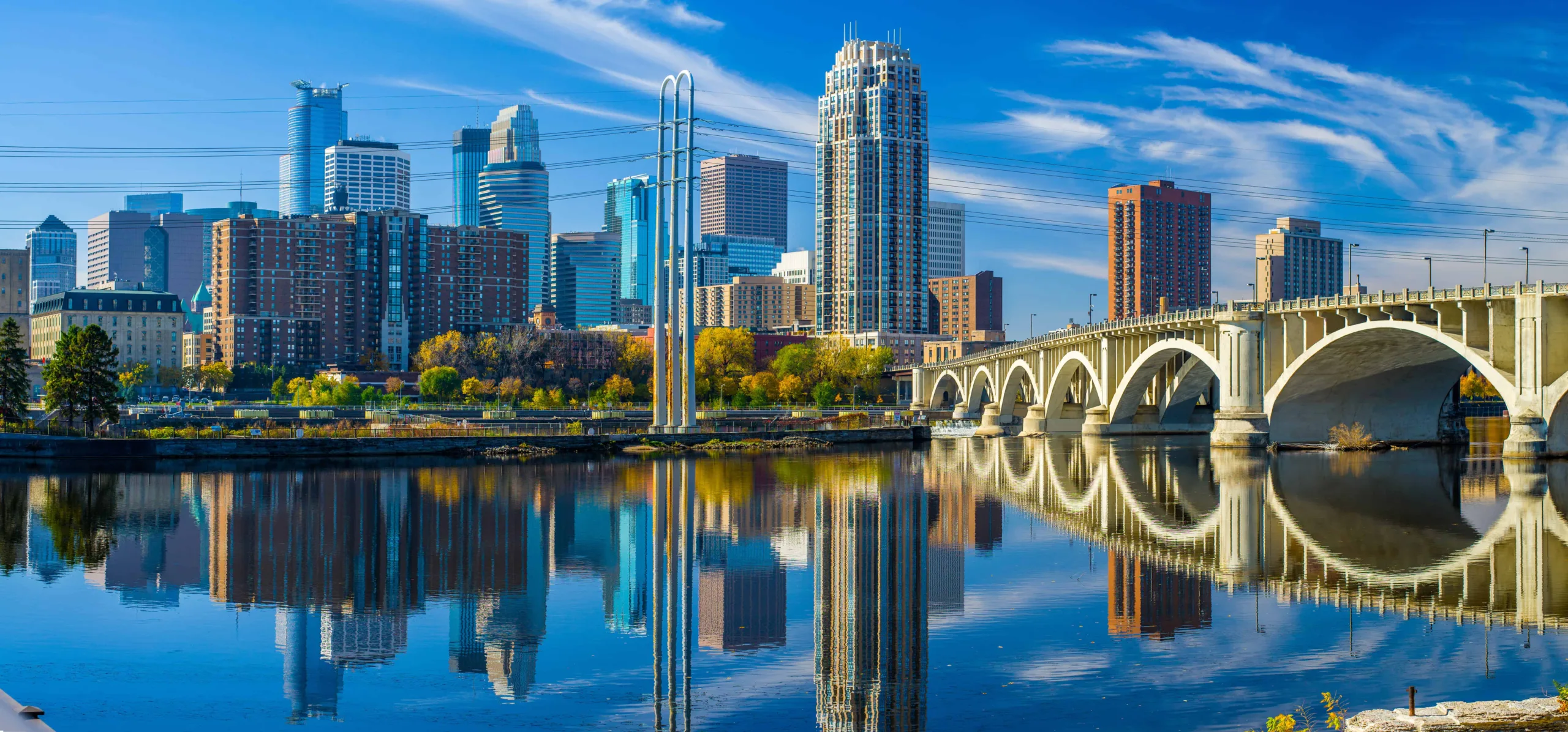 A vibrant daytime view of downtown Minneapolis, featuring the city skyline, Mississippi River, and the iconic Third Avenue Bridge, home to the University of Minnesota Medical School and its distinguished Chair of Orthopedic Surgery.